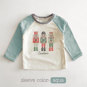 May include: A long-sleeve raglan shirt with aqua sleeves and a cream-colored body. The front features a graphic of three nutcracker figures in red, green, and grey, with the name Easton printed in black script text below the figures.