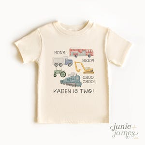 May include: Cream-colored toddler t-shirt featuring watercolor illustrations of a fire truck, dump truck, tractor, excavator, and train. Text includes "HONK!", "BEEP!", "CHOO CHOO!", and "KADEN IS TWO!" A fun shirt for a two-year-old.