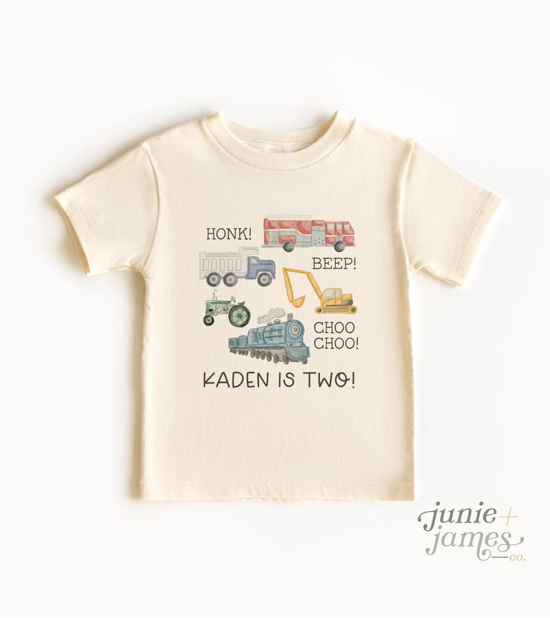 May include: Cream-colored toddler t-shirt featuring watercolor illustrations of a fire truck, dump truck, tractor, excavator, and train. Text includes "HONK!", "BEEP!", "CHOO CHOO!", and "KADEN IS TWO!" A fun shirt for a two-year-old.