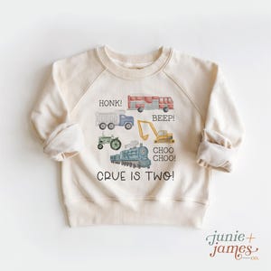 May include: Cream-colored sweatshirt with a watercolor design of vehicles, including a truck, fire truck, tractor, excavator, and train. Text on the shirt reads "HONK!", "BEEP!", "CHOO CHOO!", and "CRUE IS TWO!"