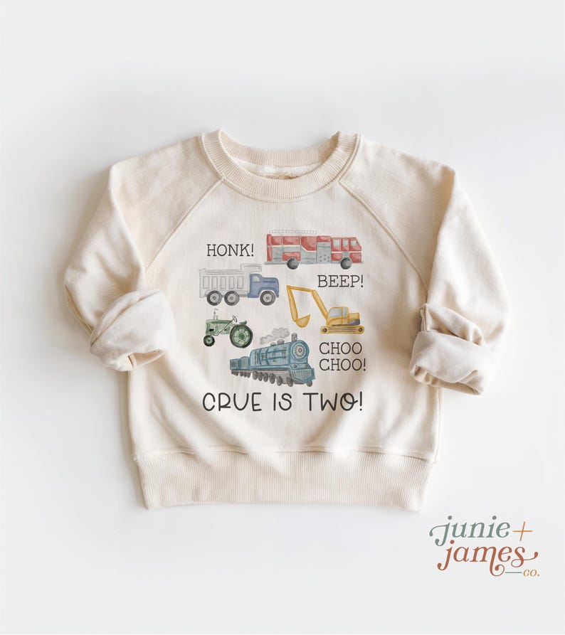 May include: Cream-colored sweatshirt with a watercolor design of vehicles, including a truck, fire truck, tractor, excavator, and train. Text on the shirt reads "HONK!", "BEEP!", "CHOO CHOO!", and "CRUE IS TWO!"