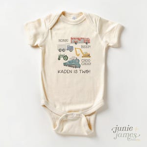 May include: Cream-colored baby onesie with watercolor illustrations of a truck, fire truck, tractor, excavator, and train. Text includes "HONK!", "BEEP!", "CHOO CHOO!", and "KADEN IS TWO!". The onesie has short sleeves and a lap shoulder neckline.