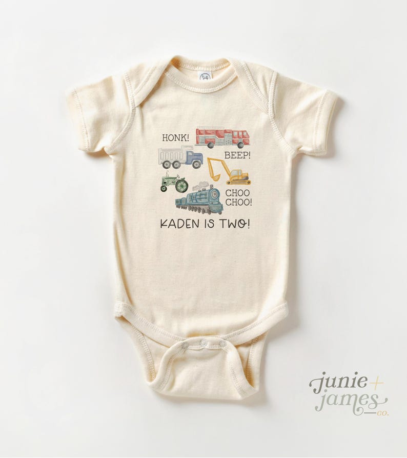 May include: Cream-colored baby onesie with watercolor illustrations of a truck, fire truck, tractor, excavator, and train. Text includes "HONK!", "BEEP!", "CHOO CHOO!", and "KADEN IS TWO!". The onesie has short sleeves and a lap shoulder neckline.