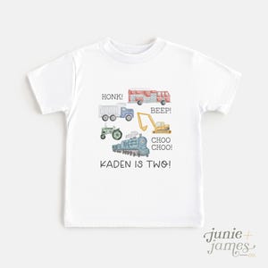 May include: White t-shirt featuring watercolor illustrations of various vehicles, including a fire truck, dump truck, tractor, excavator, and train. Text on the shirt reads "HONK!", "BEEP!", "CHOO CHOO!", and "KADEN IS TWO!"