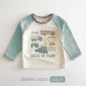May include: A long-sleeved, off-white and aqua-colored shirt with a crew neck. The shirt features illustrations of various vehicles, including a truck, fire truck, tractor, excavator, and train. Text on the shirt reads "HONK!", "BEEP!", "CHOO CHOO!", and "CRUE IS TWO!"