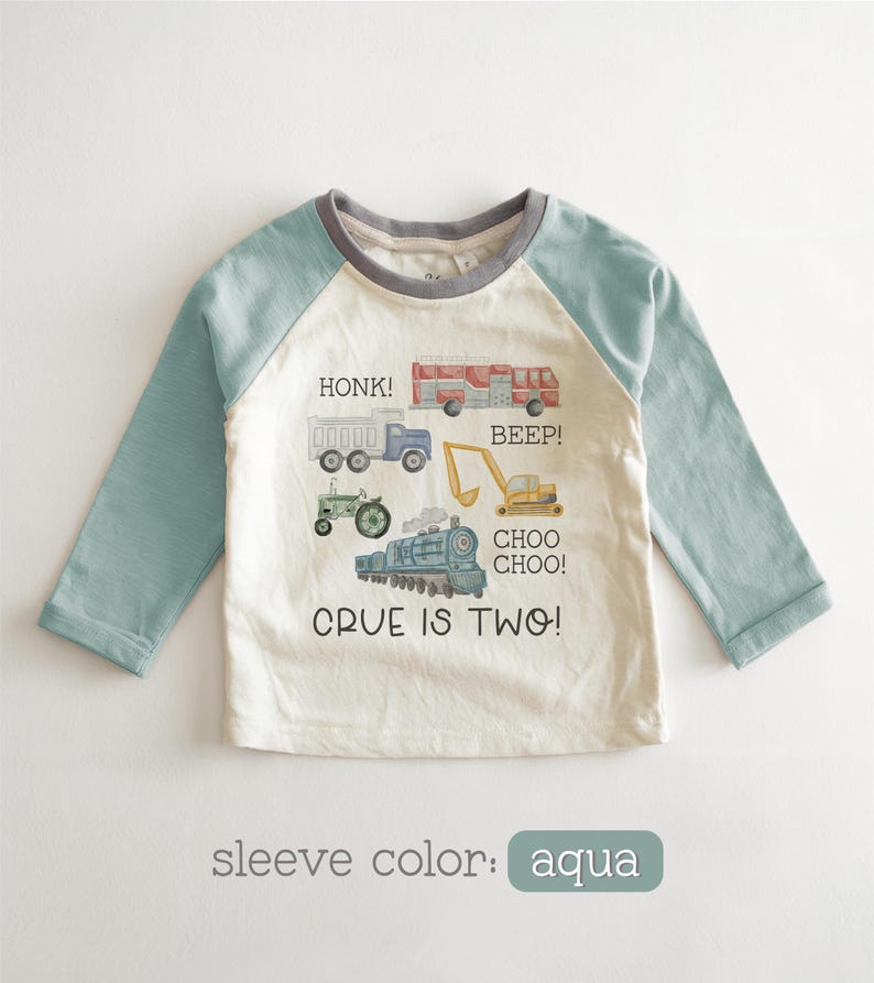 May include: A long-sleeved, off-white and aqua-colored shirt with a crew neck. The shirt features illustrations of various vehicles, including a truck, fire truck, tractor, excavator, and train. Text on the shirt reads "HONK!", "BEEP!", "CHOO CHOO!", and "CRUE IS TWO!"