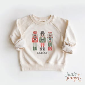 May include: A cream-colored crewneck sweatshirt featuring a graphic of three nutcracker figures in red, green, and light blue uniforms. The name Easton is printed in black script below the nutcrackers. The brand name junie + james co. is visible.