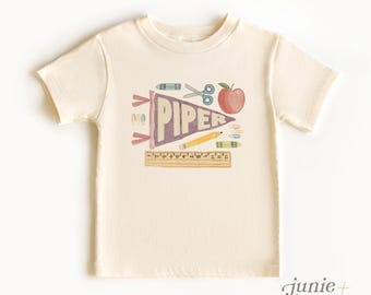 Puede incluir: Una camiseta de manga corta de color crema con un estampado gráfico de material escolar que incluye una regla, un lápiz, unas tijeras, una manzana y ceras alrededor de un banderín morado con el nombre PIPER en letras mayúsculas.