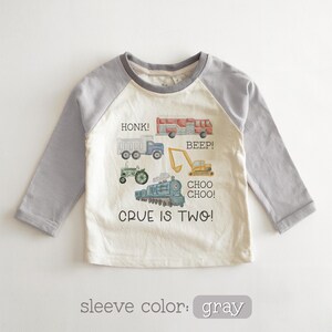 May include: A long-sleeved, raglan-style toddler shirt with a cream-colored body and gray sleeves. The shirt features colorful illustrations of vehicles, including a truck, fire truck, tractor, excavator, and train, with the text "CRUE IS TWO!"