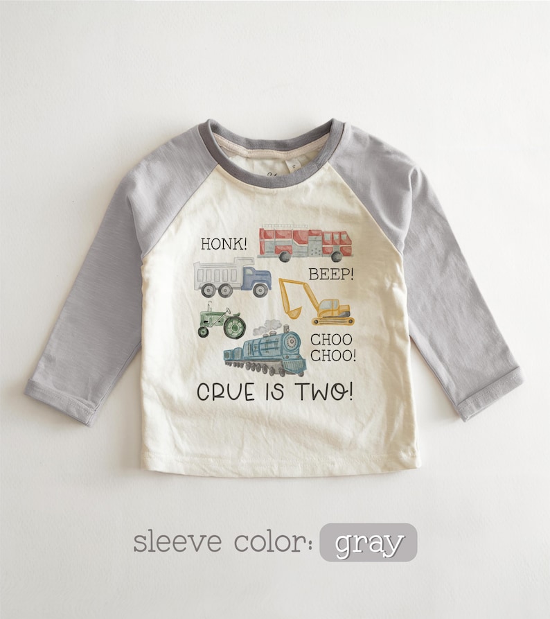 May include: A long-sleeved, raglan-style toddler shirt with a cream-colored body and gray sleeves. The shirt features colorful illustrations of vehicles, including a truck, fire truck, tractor, excavator, and train, with the text "CRUE IS TWO!"