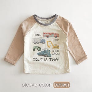 May include: A long-sleeved, off-white toddler shirt with brown sleeves and a gray neckline. The shirt features colorful illustrations of vehicles, including a truck, tractor, fire truck, and train, with the text "HONK!", "BEEP!", and "CHOO CHOO!". The text "CRUE IS TWO!" is also printed on the shirt.