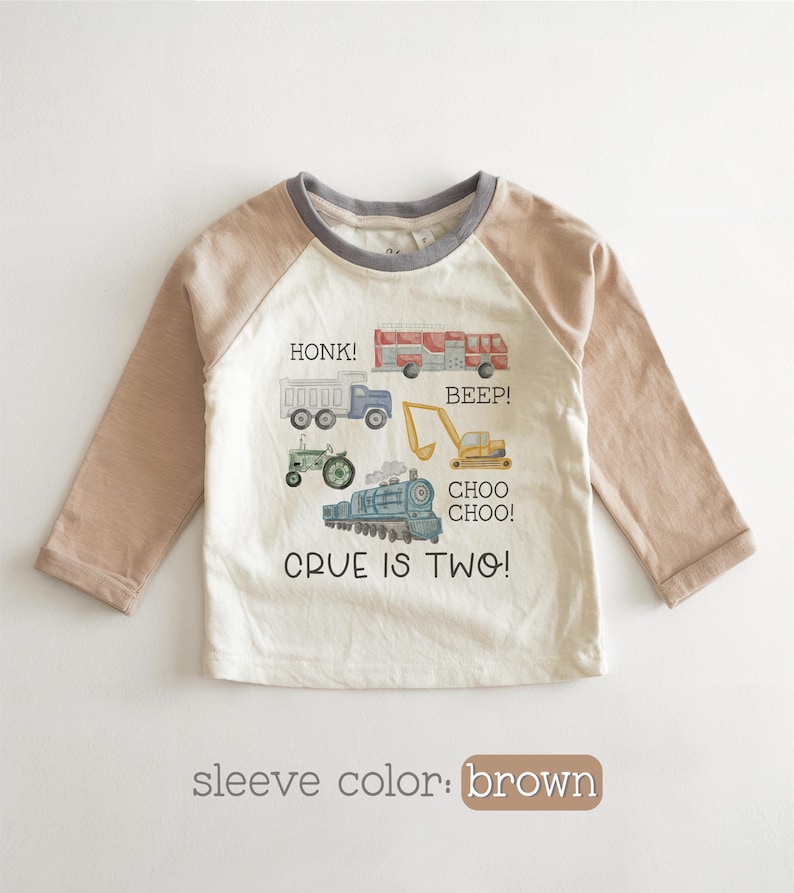 May include: A long-sleeved, off-white toddler shirt with brown sleeves and a gray neckline. The shirt features colorful illustrations of vehicles, including a truck, tractor, fire truck, and train, with the text "HONK!", "BEEP!", and "CHOO CHOO!". The text "CRUE IS TWO!" is also printed on the shirt.