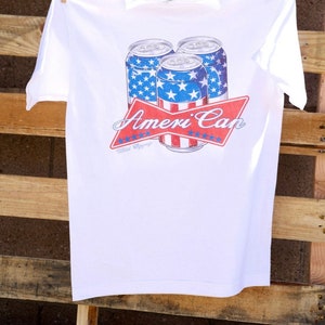 May include: White t-shirt with a graphic of three red, white, and blue beer cans with stars and stripes. The text "Ameri'Can" is printed in red with white stars on a red ribbon.