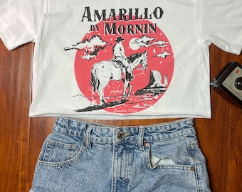 May include: White cropped t-shirt with a red, white, and black graphic of a cowboy riding a horse in front of a red sun and the text "Amarillo by Mornin'" and "Country Decor".