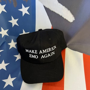May include: Black baseball cap with the text "MAKE AMERICA EMO AGAIN" in white. The hat is displayed against a backdrop of an American flag.