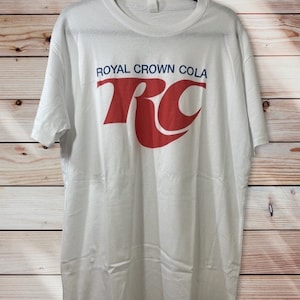 May include: White t-shirt with a red and blue "Royal Crown Cola" logo. The logo features the letters "RC" in a stylized font.