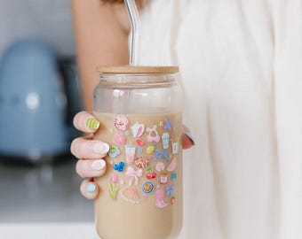 Op de afbeelding: Een herbruikbare glazen beker met een bamboe deksel en een heldere glazen rietje. De beker is bedekt met kleurrijke stickers in verschillende vormen en ontwerpen. De beker is gevuld met een lichtbruine drank.