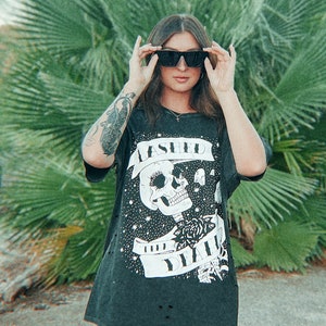 以下が含まれることがあります： 黒いTシャツに頭蓋骨のグラフィックと「Assault and Battery」の文字、黒い太もも丈のブーツを履いた女性。彼女はヤシの木の前に立っている。