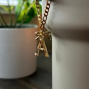 Pode incluir: Um colar de corrente de ouro com um pingente de letra A de ouro e um laço. O pingente é feito para parecer um galho.