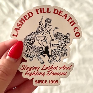 May include: White sticker with red text that says "Lashed Till Death Co. Slaying Lashes And Fighting Demons Since 1995". The sticker features a black and white illustration of a devil with a pitchfork standing on two tigers.