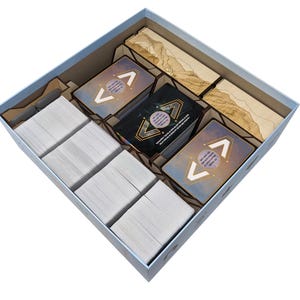 Op de afbeelding: Een lichtblauwe speldoos gevuld met georganiseerde spelonderdelen. De doos bevat stapels witte kaarten, spelkaarten met geometrische ontwerpen en houten verdelers. De speelkaarten hebben een hemels thema.