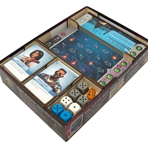 May include: A board game box organiser containing two stacks of character cards featuring Lucia Ortega and Clinton Harper, a dark blue game board with white grid lines, and several six-sided dice in blue, white, grey, and orange colours.