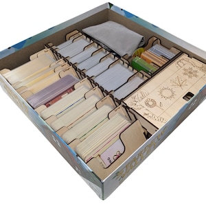 May include: Wooden organiser insert for the board game "The Gardens of Babylon". The insert has compartments for cards, tokens, and a cloth bag. The insert is made of light brown wood and has a natural finish.