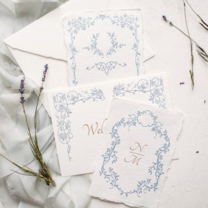 May include: Four white cards with blue floral designs. The cards have different designs, including a corner design, a full border design, and a monogram design. The monogram design includes the initials "N" and "M".