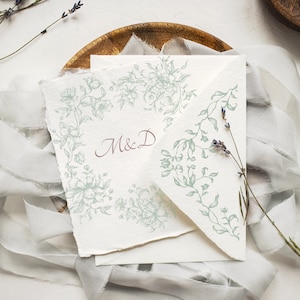 May include: A white card with a green floral design and the initials "M&D" in maroon ink. The card is on a wooden plate with a white fabric overlay.