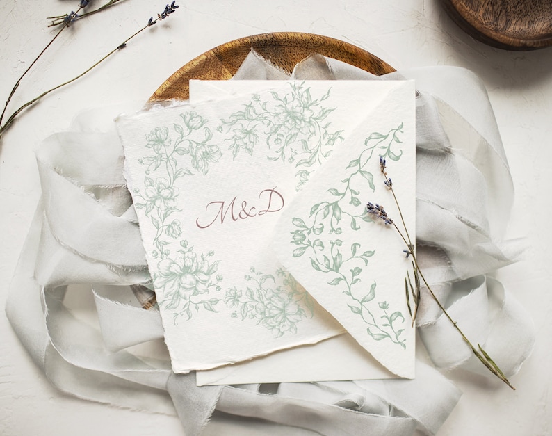 May include: A white card with a green floral design and the initials "M&D" in maroon ink. The card is on a wooden plate with a white fabric overlay.