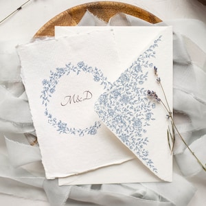 May include: A white card with a blue floral design on the envelope flap. The card has a blue floral wreath with the initials "M&D" in the center.