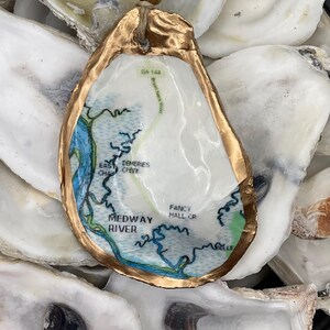 May include: A gold-rimmed oyster shell with a map of the Midway River area in Georgia. The map shows the river, creeks, and roads. The oyster shell is surrounded by other oyster shells.