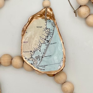 May include: An oyster shell ornament with a gold-painted edge, featuring a map of Barnegat, Ship Bottom, and Beach Haven. The shell is suspended by a jute twine string with a single light blue bead and natural wooden beads on either side.