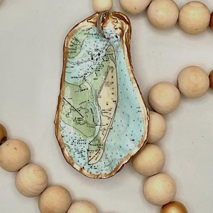 May include: A decorative oyster shell ornament featuring a decoupage map of Jekyll Island with gold-painted edges, suspended from a string of natural wooden beads and a single light blue bead, creating a coastal-themed hanging accessory.