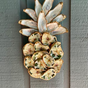 May include: A decorative wall hanging shaped like a pineapple, constructed from individual oyster shells. The top leaves are iridescent white shells, while the base consists of shells decoupaged with white floral patterns, all featuring gold-painted edges.