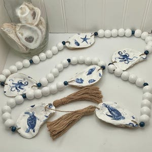May include: White beaded garland with painted oyster shells featuring blue sea creatures and nautical designs. Includes two brown tassels. A glass jar with oyster shells is in the background.