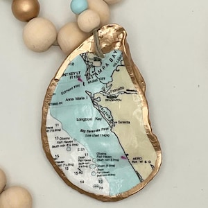 May include: An oyster shell pendant featuring a map of Tampa Bay, Florida, with gold-painted edges. The pendant is strung with natural wooden beads and a small blue bead. A unique coastal-themed accessory.