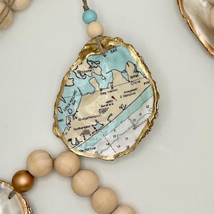 May include: A decorative garland featuring natural wooden beads in matte and metallic gold finishes, interspersed with oyster shells. Each shell is edged in gold paint and features a decoupage map of the Hamptons, including Sag Harbor and Southampton.