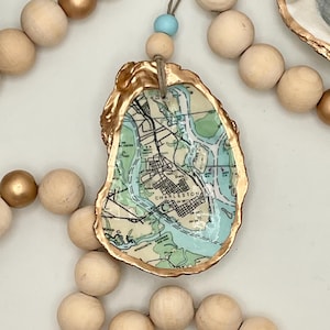 May include: An oyster shell pendant featuring a map of Charleston, South Carolina, with a gold-painted edge. The pendant hangs from a natural cord with a light blue bead, surrounded by wooden beads.