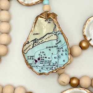 May include: A decorative garland featuring natural oyster shells with gold-painted edges and a decoupage map of Kiawah Island, Seabrook Island, and Johns Island. The shells are strung together with wooden beads in natural and metallic gold finishes.
