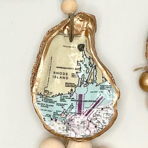 May include: A handcrafted oyster shell ornament featuring a map of Rhode Island. The shell has a gold-painted edge and is strung with natural wooden beads and a blue accent bead. The map details the Narragansett Bay and Block Island Sound.