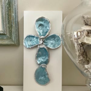 May include: A decorative cross made from five blue oyster shells with silver-colored edges, mounted on a white rectangular base. The shells have a glossy finish. The artwork is displayed on a white surface.