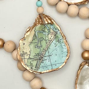 May include: A decorative garland featuring natural wooden beads, metallic gold beads, and a light blue bead. The garland includes oyster shells with gold-painted edges, each decoupaged with a nautical map showing Sea Island and Village Creek.