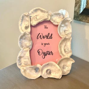 May include: A decorative picture frame with a pink background and the phrase "The World is your Oyster" in black script. The frame is made of oyster shells, creating a unique coastal aesthetic.