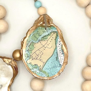 May include: A decorative garland featuring natural wooden beads and oyster shells with gold-painted edges. The center shell displays a map of Daufuskie Island with green land and blue water details. A single light blue bead accents the twine cord.
