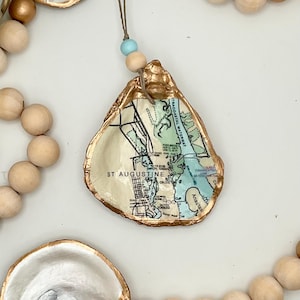 May include: A decorative oyster shell ornament featuring a decoupage map of St. Augustine, Florida, with gold-painted edges. The shell hangs from a string adorned with natural wooden beads and a single light blue bead, creating a coastal garland.