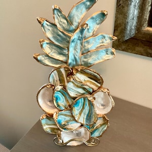 May include: A decorative pineapple sculpture constructed from individual oyster shells. The top leaves are painted in a metallic turquoise, while the base consists of shells featuring marbled swirls of white, gold, and turquoise, all edged in gold.