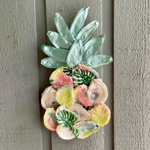 May include: A decorative wall hanging shaped like a pineapple, constructed from individual oyster shells. The top leaves are painted a solid light mint green, while the base consists of shells decorated with colorful tropical leaf patterns in pink, yellow, and green.