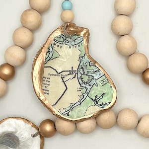 May include: A decorative garland featuring natural wooden beads in matte and metallic gold finishes, interspersed with oyster shells. One shell is decoupaged with a nautical map of the May River and Palmetto Bluff, featuring gold-painted edges.