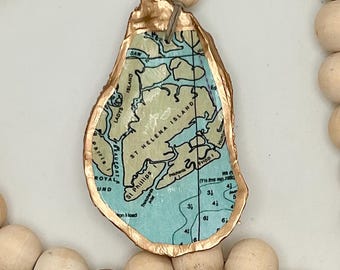 Può includere: Un ciondolo decorativo a forma di conchiglia di ostrica con una mappa di St. Helena Island, Carolina del Sud, con bordi dipinti d'oro. Il ciondolo è infilato con perline di legno e una perlina di accento azzurro. La mappa include nomi di luoghi come "Fripp I".